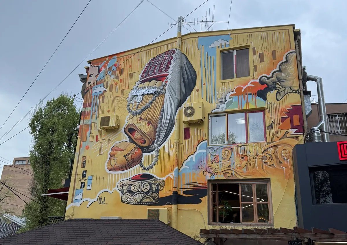 Mural of a woman in traditional Armenian dress with silver jewellery and clay vessels covering the wall of a yellow building