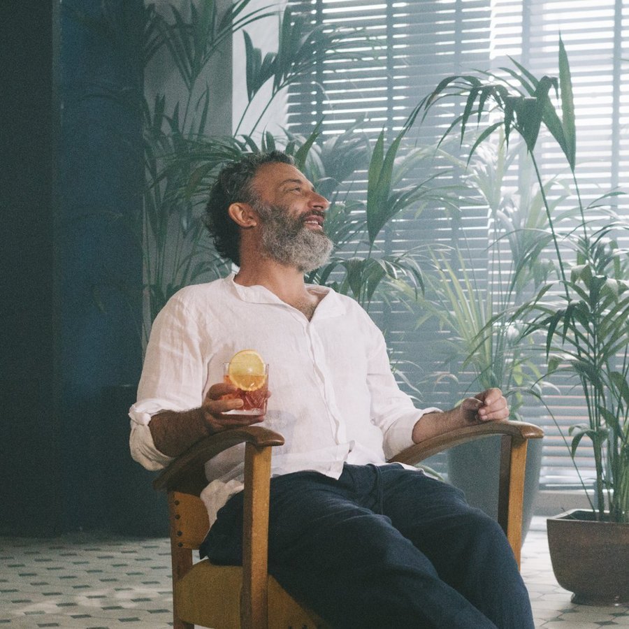 Igor Gurovich seated in a wooden chair with a cocktail, smiling, surrounded by green plants