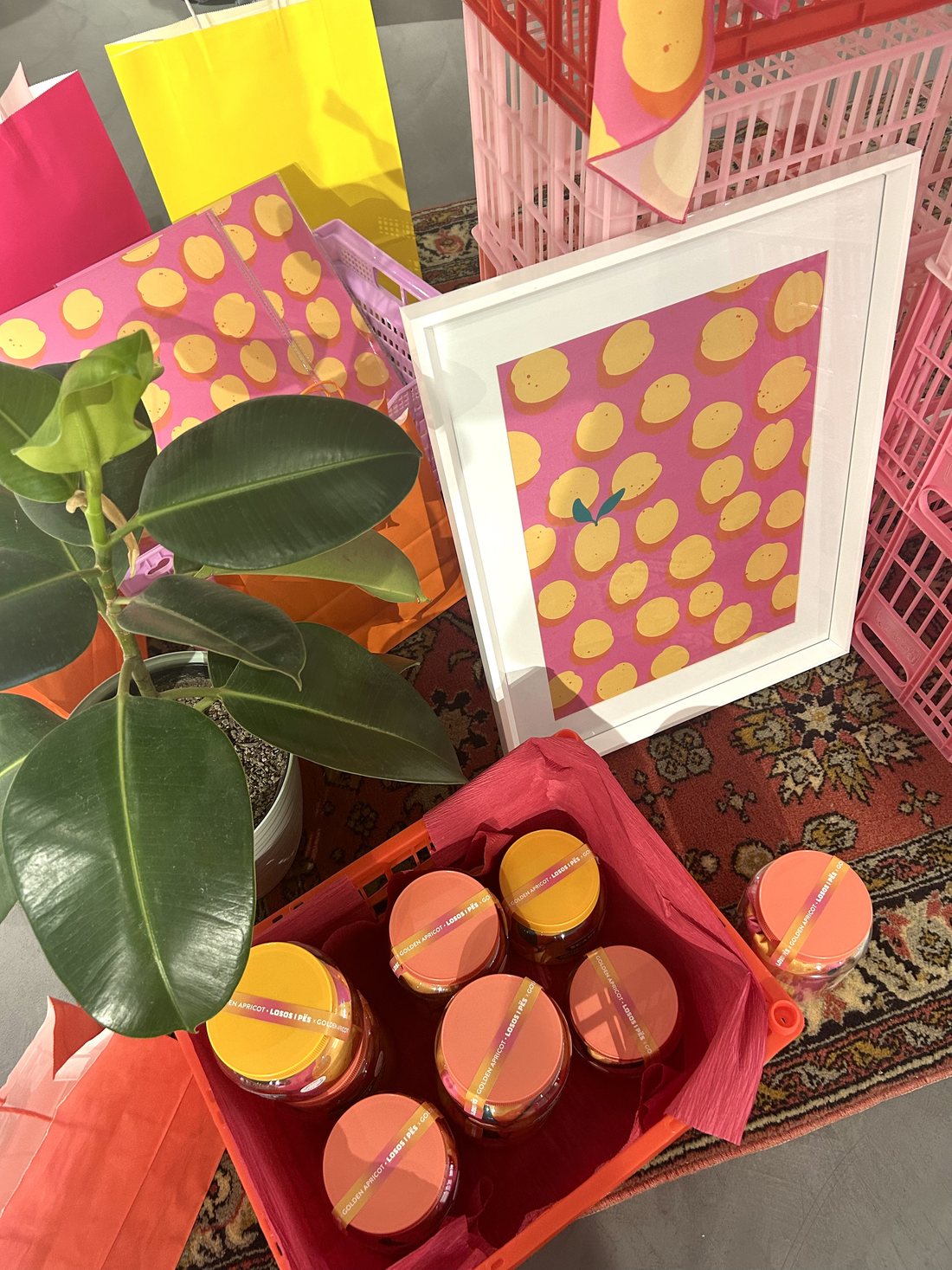 A framed print of golden apricots scattered across a bright pink background, surrounded by matching tote bags and jars